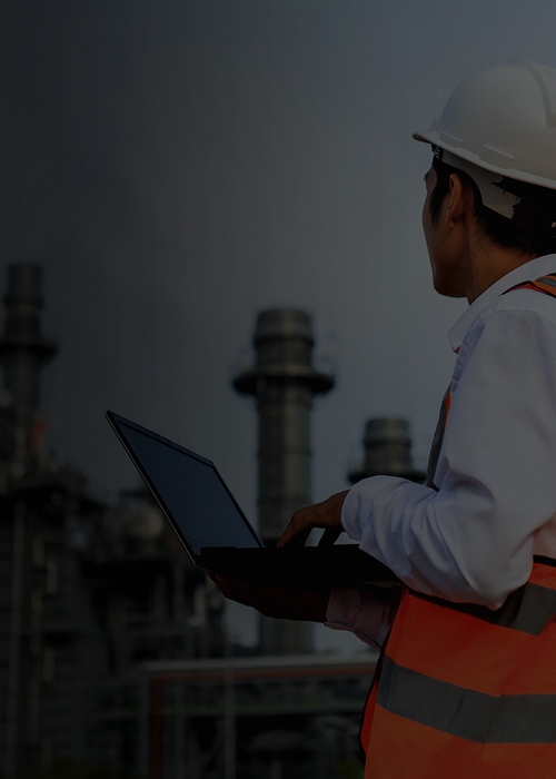 Worker with protective gear using computer in front of oil and gas industry